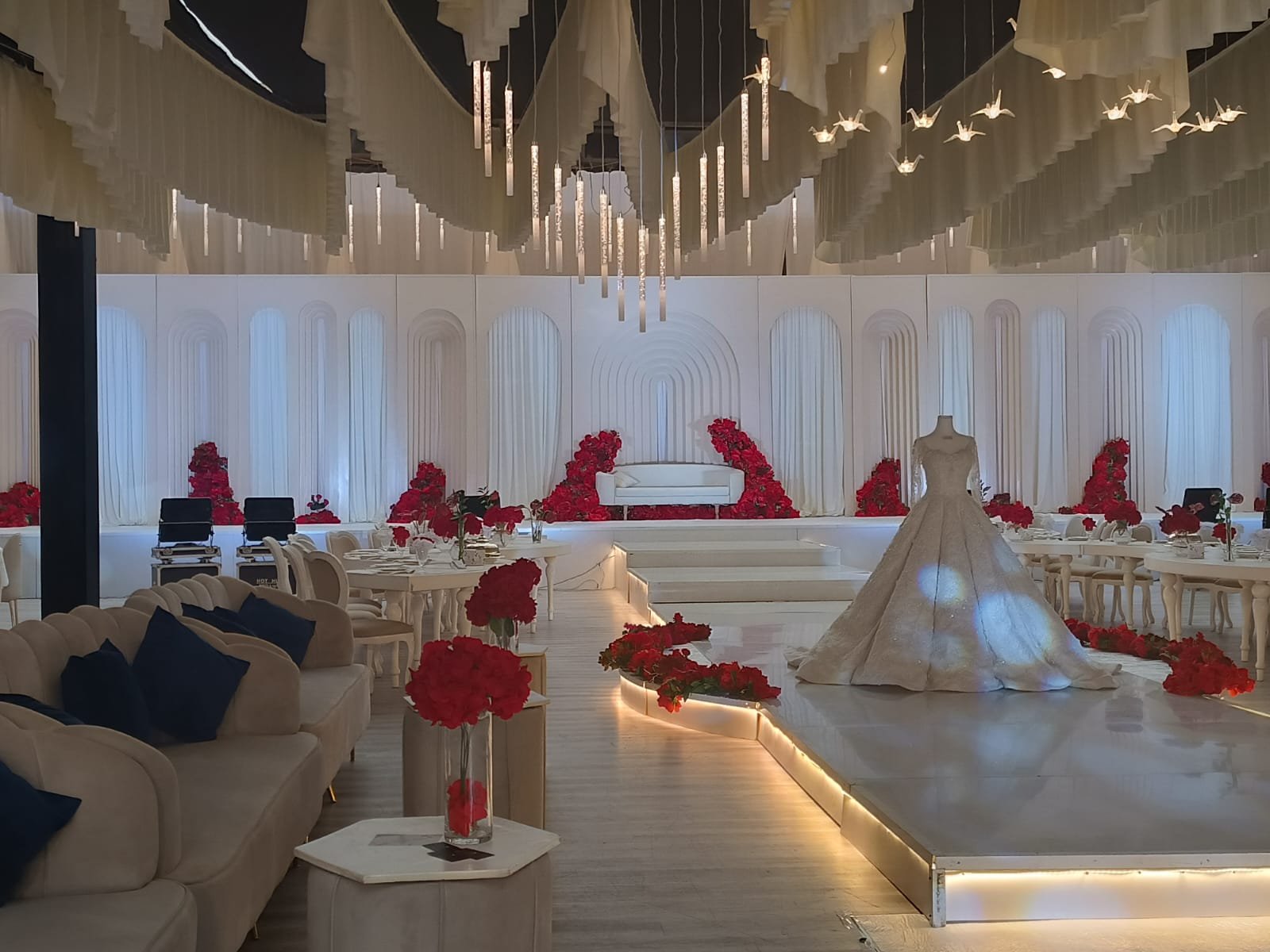 ديليزيا  العين - ديكورات قاعات أفراح
                                منصة زفاف فاخرة في قاعة أفراح واسعة. المنصة مرتفعة قليلاً عن مستوى الأرضية ومزينة بالورود الحمراء بشكل كثيف.  الإضاءة هادئة وطبيعية، مما يخلق جوًا رومانسيًا ودافئًا